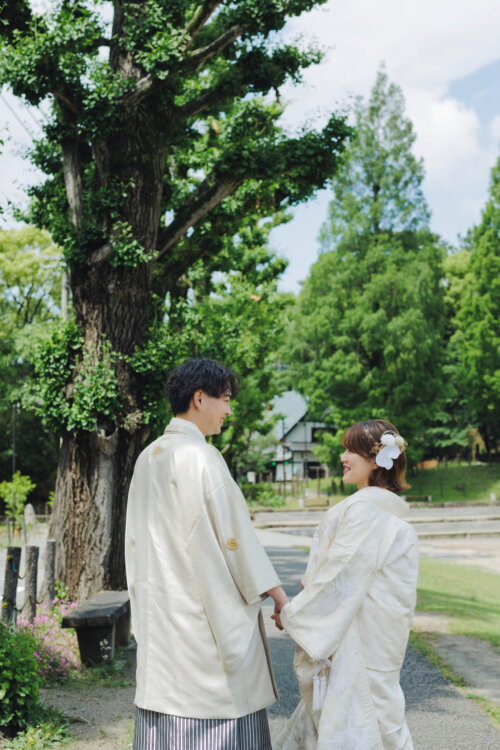 大きな木の下で向かい合って手を取る、白紋付袴の新郎と白無垢の花嫁。新緑の公園で撮るロケーションフォト
