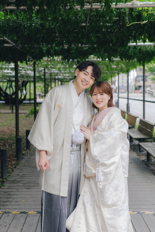 藤棚の下の木道で寄り添って笑顔を見せる、白紋付袴の新郎と白無垢の花嫁。仲の良さが伝わる結婚写真