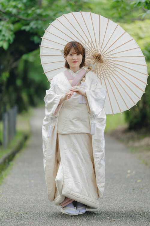 木陰の小道で和傘を差して立つ、ピンクの掛下を合わせた白無垢SF105の花嫁。和装前撮りの全身ショット
