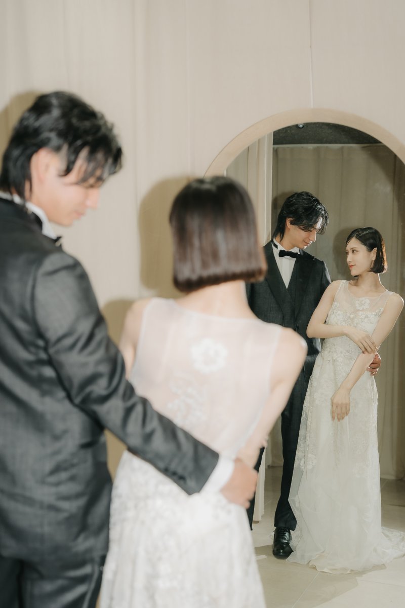 レースドレス姿の新婦とダークスーツの新郎がアーチ型の鏡越しに見つめ合う、ナチュラルな光の中で