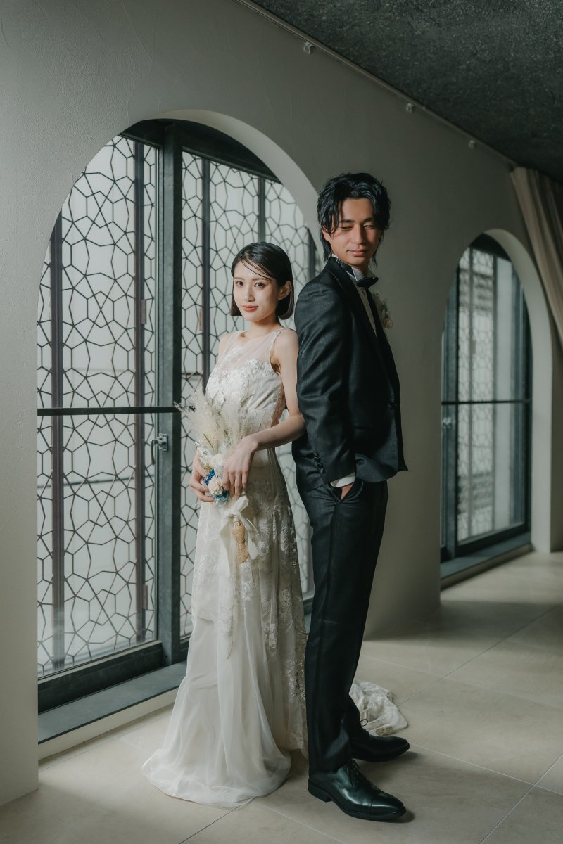 黒いタキシード姿の新郎とレースドレス姿の新婦が背中合わせに立つ、アーチ窓のあるスタジオにて