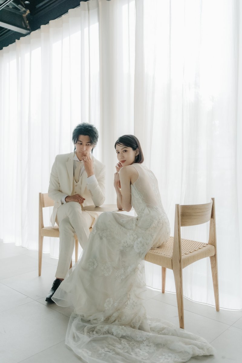 アイボリーのタキシード姿の新郎とレース刺繍のドレス姿の新婦が丸テーブルを囲む、白いカーテン越しの柔らかな光の中で