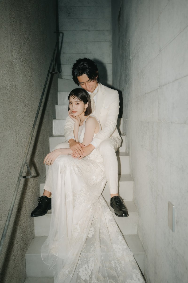 白いタキシード姿の新郎とレースドレス姿の新婦が寄り添う、コンクリート階段に腰掛けた後ろ姿