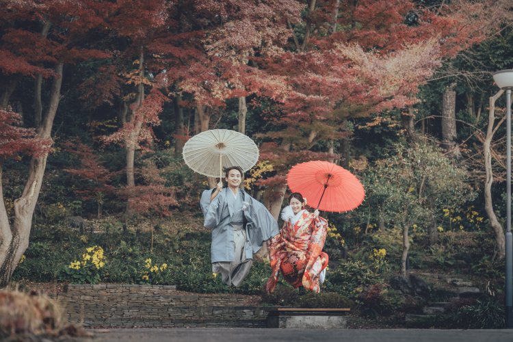 紅葉の木々の下で白い和傘と赤い和傘を差す水色の羽織袴の新郎と赤い色打掛の花嫁を引きでとらえた一枚。秋の日本庭園で撮る和装ロケーションフォト