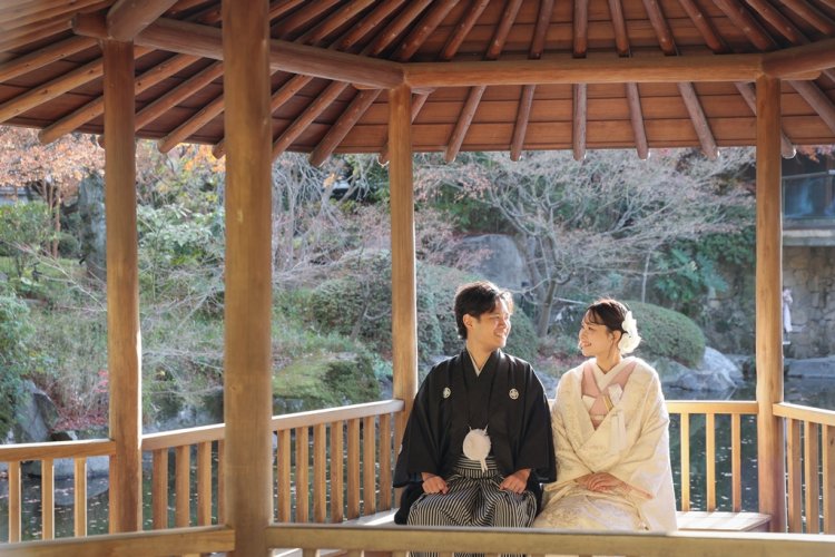 池を望む東屋のベンチに座って見つめ合う黒紋付袴の新郎と白い色打掛の花嫁を引きでとらえた一枚。秋の目白庭園で撮る和装前撮り