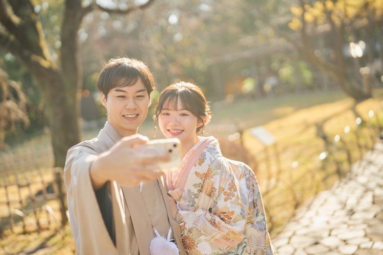 紅葉の庭園でスマートフォンを掲げてセルフィーを撮るベージュの羽織袴の新郎と花柄の色打掛の花嫁。平成庭園で撮る和装前撮りの遊び心のあるカット