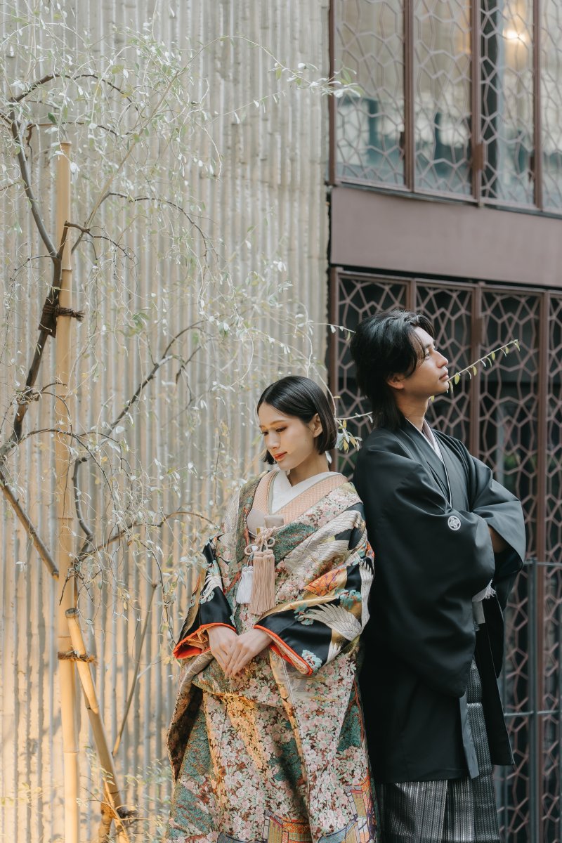 黒紋付袴姿の新郎と華やかな色打掛姿の新婦が背中合わせに佇む、格子窓のアート建築を背景に