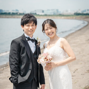 弧を描く砂浜を背に、ブーケを持って並んで笑うタキシードの新郎とレースのドレスの花嫁。葛西臨海公園で撮る前撮り