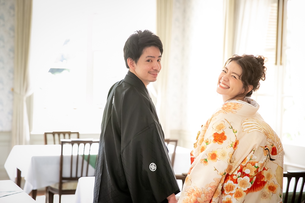 洋館のレストランで振り返って笑う黒紋付の新郎と、橙色の花柄の色打掛の花嫁。明るい光に包まれた和装前撮り