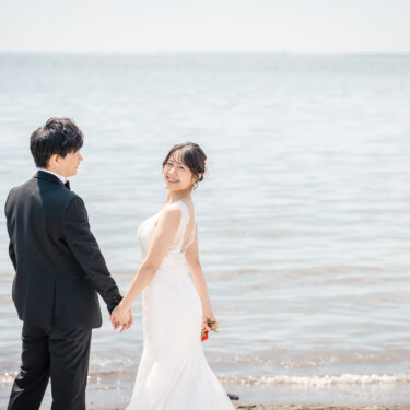 水平線に向かって手をつなぐ新郎と、振り返って笑顔を見せるマーメイドドレスの花嫁。葛西海浜公園のビーチで撮るウェディングフォト