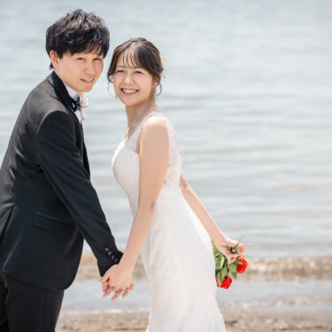 海を背に手をつなぎ、並んでカメラに顔を寄せるタキシードの新郎と花嫁。赤いバラのブーケがアクセントの前撮り