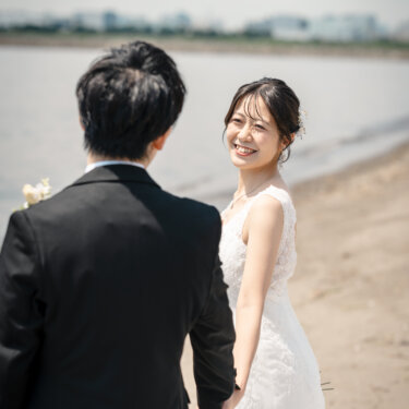砂浜で手をつないで歩きながら、振り返って笑う花嫁と新郎の後ろ姿。葛西の人工なぎさで撮るフォトウェディング