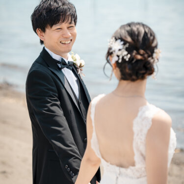 波打ち際で手をつなぎ、花嫁を見つめて笑う新郎。背中が開いたレースのウェディングドレスとヘアアレンジが映える前撮り