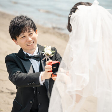 砂浜にひざまずき、赤いリングケースを開いて花嫁を見上げる新郎。ベール越しに撮ったプロポーズシーンの前撮り