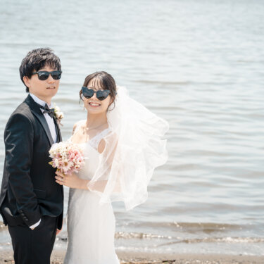海辺でサングラスをかけてポーズを決めるタキシードの新郎とブーケを持つ花嫁。ベールが風に舞う夏らしい前撮り