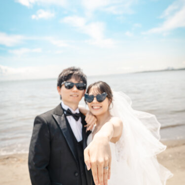サングラス姿の新郎新婦が、花嫁の指輪を見せるように手を差し出す海辺のワンシーン。おしゃれなポーズのウェディングフォト