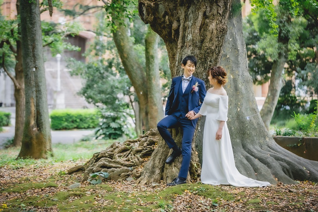 大木の根元で見つめ合うネイビーのスーツの新郎とオフショルダーのウェディングドレスの花嫁。日比谷公園の大木を背景にした洋装前撮り