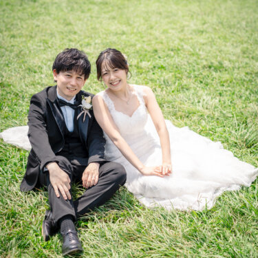 広い芝生に並んで座り、カメラに笑顔を向けるタキシードの新郎とウェディングドレスの花嫁。公園ロケならではの開放的な前撮り