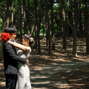 松林の小道で、赤いバラのブーケを手に新郎の首に腕を回してハグする花嫁。葛西臨海公園の林で撮る前撮り
