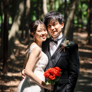 木漏れ日の林の中で、赤いバラのブーケを持って頬を寄せ合う新郎と花嫁。自然な笑顔が残るウェディングフォト