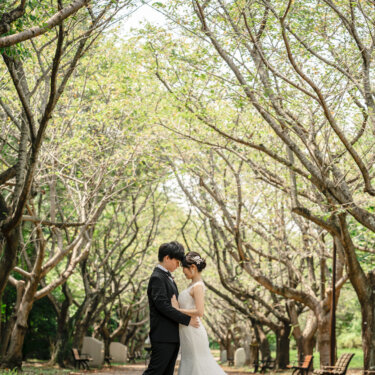 枝を広げた木々のトンネルの下で、おでこを寄せ合う新郎新婦。緑に包まれた公園で撮る洋装前撮り
