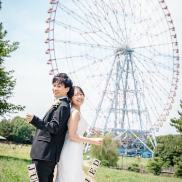 葛西臨海公園の大観覧車の下で、背中合わせになりJUST MARRIEDのガーランドを掲げる新郎新婦。青空が広がる洋装ロケーションフォト