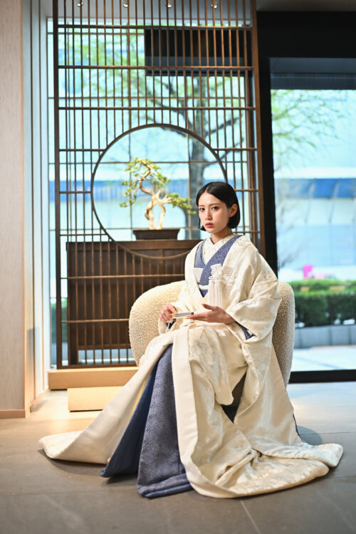 丸窓の衝立と盆栽を背に、白地の打掛と藍の掛下をまとって椅子に腰かける花嫁。和モダンなスタジオでの和装前撮り