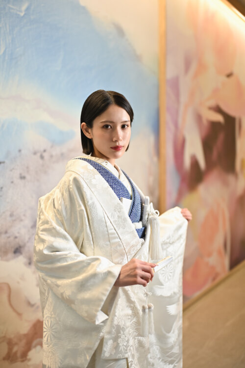 淡い色彩のアートウォールの前で末広を手にした白無垢の花嫁。藍の掛下と白の打掛が凛とした和装フォトウェディング