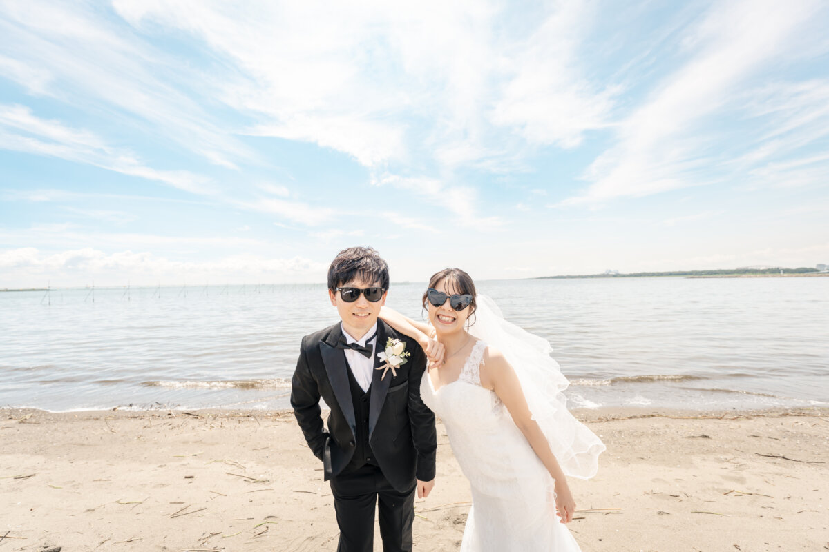青空と遠浅の海を背景に、サングラスで笑う新郎とベールをなびかせる花嫁。夏の海で撮るおしゃれな結婚写真