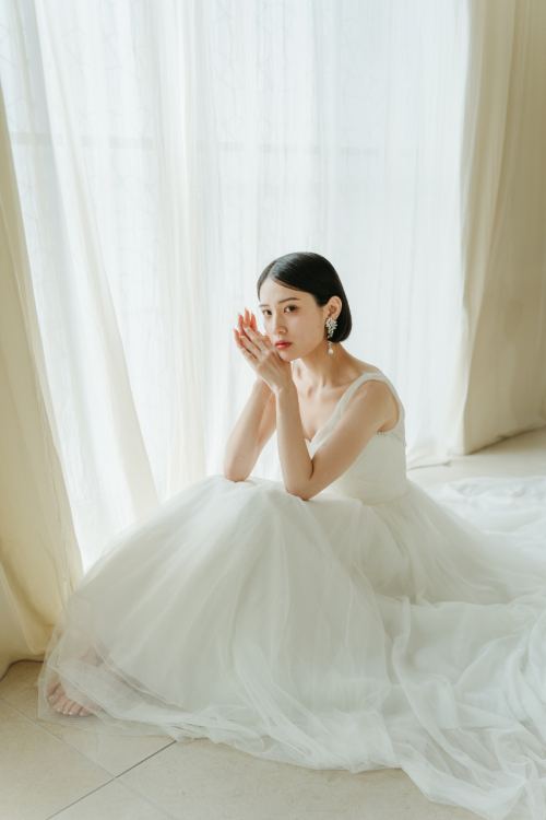 白いカーテンの前でチュールのスカートを広げて座る花嫁。逆光がドレスを透かすウェディングドレスのカタログカット