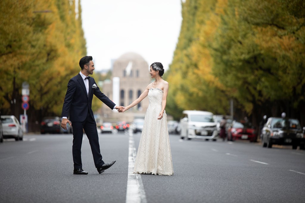 外苑いちょう並木の道の中央を手をつないで歩く、ウェディングドレスの花嫁とネイビーのタキシードの新郎