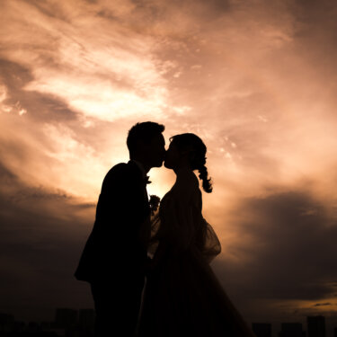 燃えるような夕焼け雲を背景にキスを交わす新郎新婦のシルエット。マジックアワーの洋装ロケーションフォト