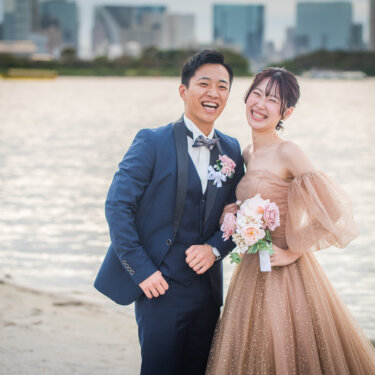 お台場海浜公園の海辺で、ブーケを持って満面の笑顔を見せるタキシードの新郎とカラードレスの花嫁の洋装前撮り