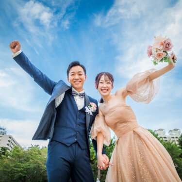 青空の下でブーケを掲げ、こぶしを突き上げて喜ぶタキシードの新郎とカラードレスの花嫁をローアングルで写した結婚写真