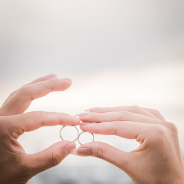 指先でつまんだふたつの結婚指輪を重ねて輪をつくる手元のアップ。海辺で撮る前撮りの指輪カット