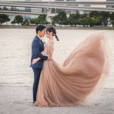 お台場海浜公園の砂浜で風になびくチュールのカラードレスの花嫁を抱き寄せ、おでこを合わせる新郎の前撮り