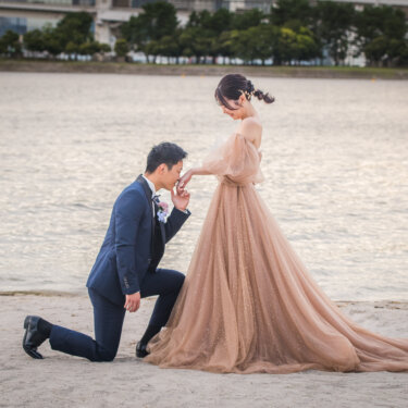 砂浜にひざまずき、カラードレスの花嫁の手の甲にキスをする新郎。プロポーズのようなポーズの洋装前撮り
