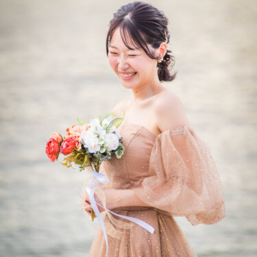 オレンジと白の花のブーケを持ち、うつむいて笑うパフスリーブのカラードレスの花嫁のソロカット。海辺の前撮り