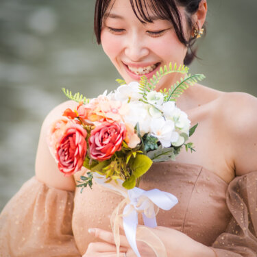 ブーケを胸元に抱え、はにかんだ笑顔を見せる花嫁のアップ。ベージュのカラードレスとヘアアレンジが映える結婚写真