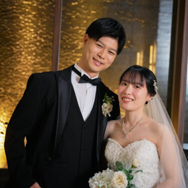 ブーケを持つ花嫁と肩を寄せて笑顔を向けるタキシードの新郎。金色の光の壁の前で撮るチャペルフォトの定番ポーズ
