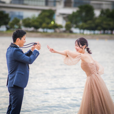 水辺でカメラを構える新郎と、両手を広げておどけるカラードレスの花嫁。お台場で撮る遊び心ある洋装前撮り