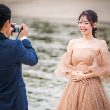 新郎がカメラで撮影するのに合わせ、海辺で笑顔を向けるカラードレスの花嫁。自然体の表情を写したフォトウェディング