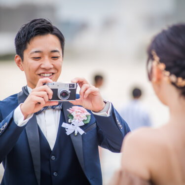 フィルムカメラを構えて花嫁を撮る笑顔のタキシードの新郎。ゴールドの髪飾りが映える花嫁の後ろ姿も入った前撮り
