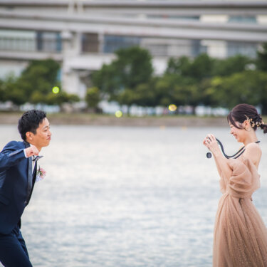 海辺でカメラを向け合うタキシードの新郎とカラードレスの花嫁。お台場海浜公園で撮る楽しい洋装ロケーションフォト