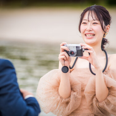 首からストラップを下げ、カメラで新郎を撮る笑顔のカラードレスの花嫁。撮り合いポーズの洋装前撮り