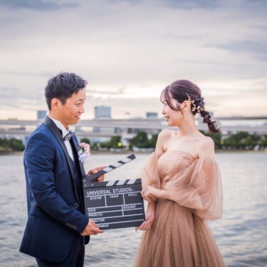 お台場の海辺でカチンコを手に見つめ合うタキシードの新郎とカラードレスの花嫁。映画風の洋装前撮り