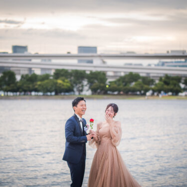 一輪の赤いバラを差し出す新郎と、口元に手を当てて驚くカラードレスの花嫁。海辺でのプロポーズ演出のフォトウェディング