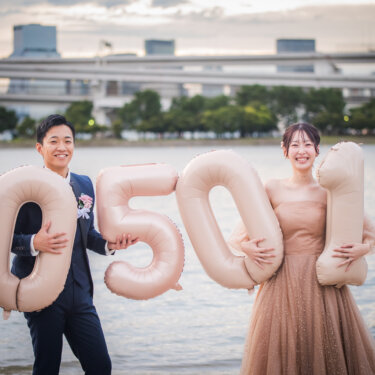 記念日の日付の数字バルーンを並べて持つタキシードの新郎とカラードレスの花嫁。お台場海浜公園の前撮り