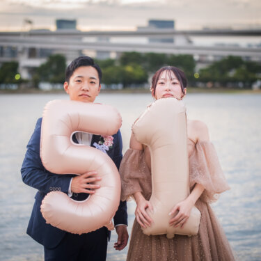 大きな数字バルーンを胸元に抱えて真顔で並ぶ新郎新婦。記念日の日付を入れた遊び心ある洋装前撮り