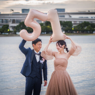 数字バルーンを頭上に掲げて並ぶタキシードの新郎とカラードレスの花嫁。海辺で撮るおしゃれなフォトウェディング